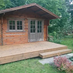 Mała drewniana chatka z dwuskrzydłowymi drzwiami i oknem, z przylegającym tarasem z desek, otoczona zielenią ogrodu.