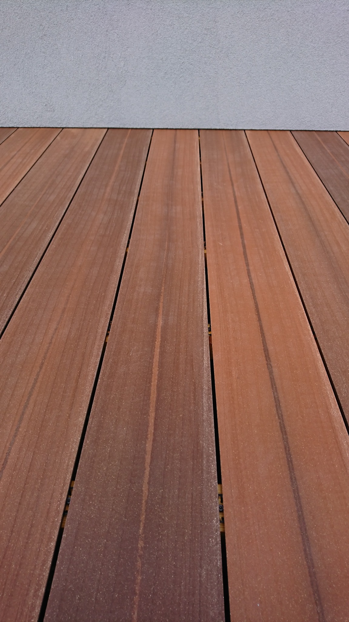 Nowoczesny taras z desek kompozytowych w odcieniach brązu, ułożonych równolegle do szarej ściany budynku.