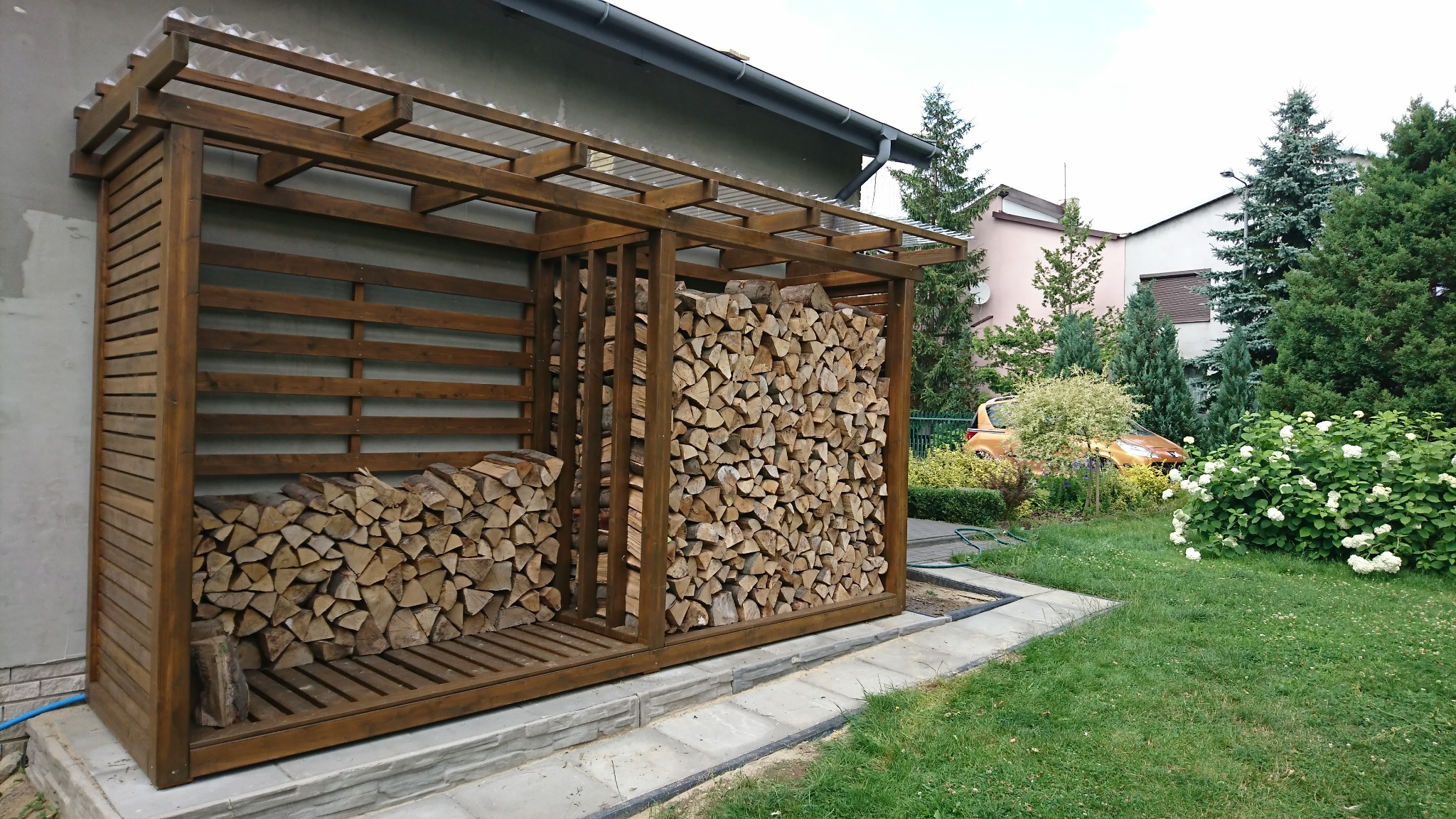 Drewniana konstrukcja z przezroczystym zadaszeniem, służąca jako skład drewna opałowego, wypełniona pociętymi kawałkami drewna, umieszczona przy ścianie budynku w ogrodzie.