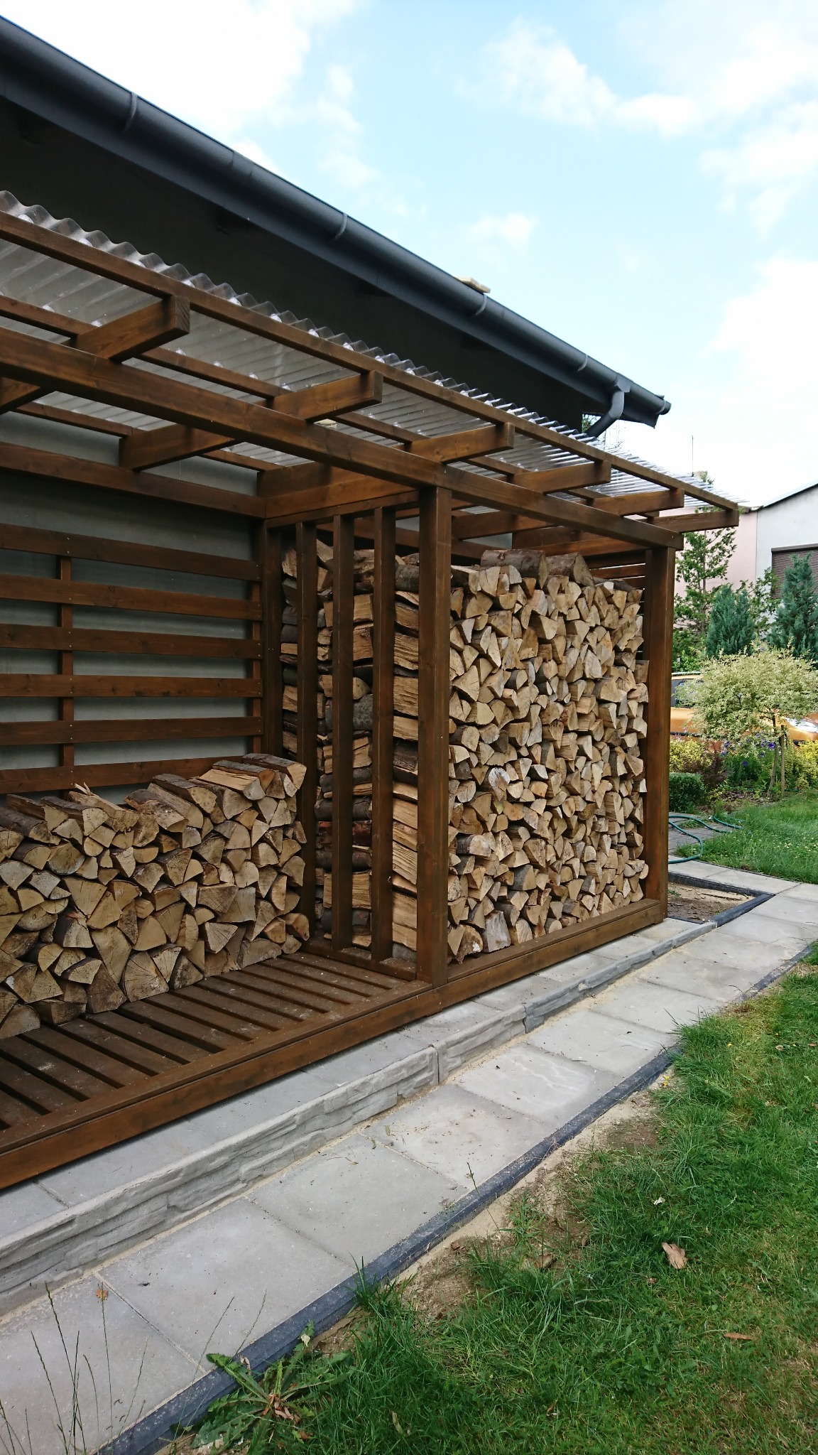 Zadaszone, drewniane miejsce do składowania drewna opałowego, z wypełnionymi dwoma przegrodami i częściowo trzecią, ustawione na zewnątrz na kostce brukowej z widocznym trawnikiem i budynkiem w tle.