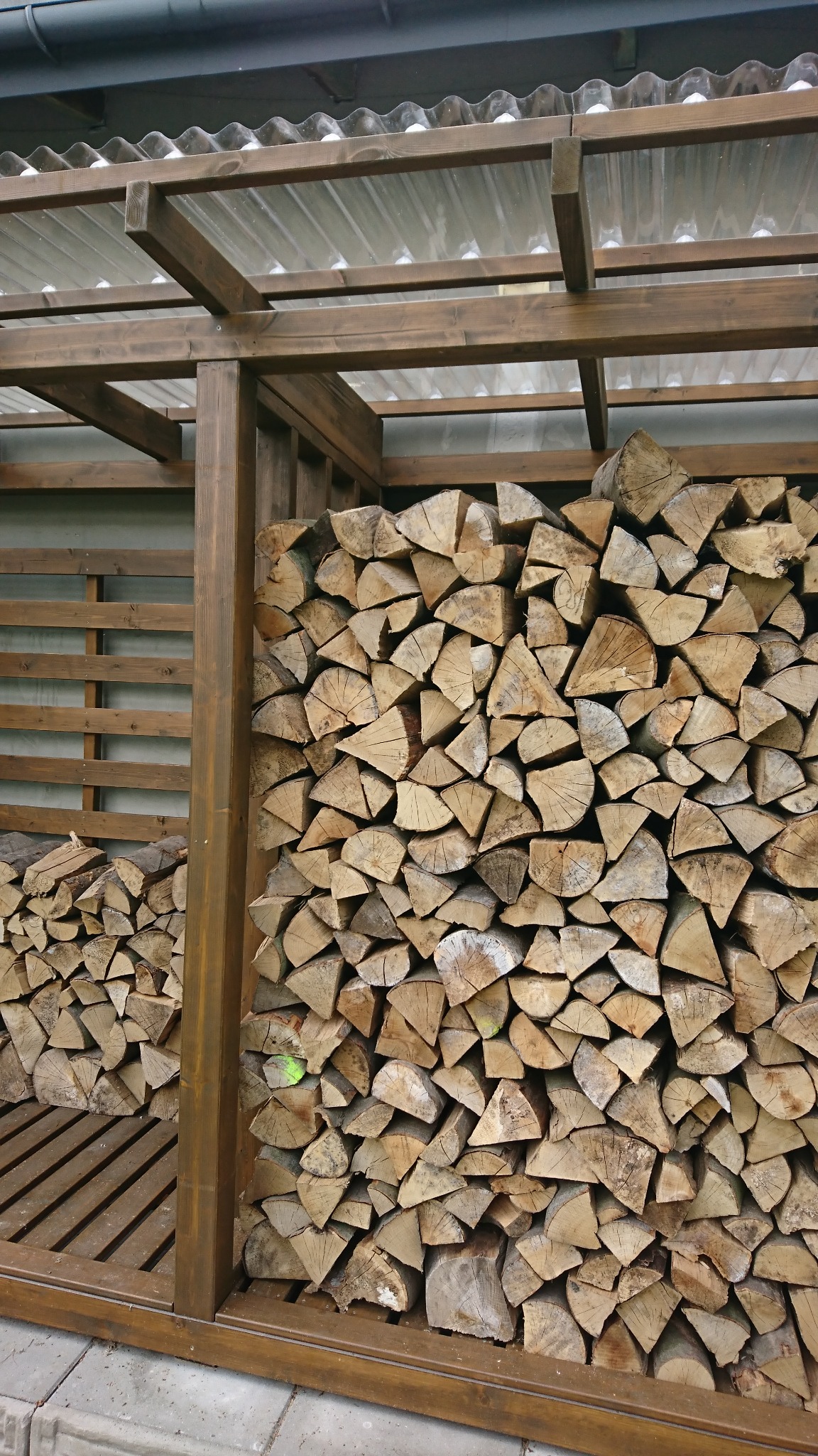Drewniana konstrukcja z zadaszeniem z tworzywa sztucznego, wypełniona pociętym drewnem opałowym, ułożonym w regularny stos.