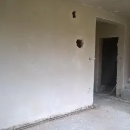 Wnętrze po nałożeniu gładzi gipsowej, widoczne przygotowane otwory na gniazdka elektryczne i puszki instalacyjne, surowy beton schodów oraz przejście do innego pomieszczenia w stanie deweloperskim.