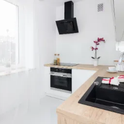 Jasna kuchnia z drewnianym blatem, czarnym zlewem i nowoczesną armaturą, widok na okno z białymi zasłonami, czarny okap nad płytą grzewczą i minimalistyczny wystrój.