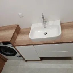 Łazienka z umywalką nablatową na szafce z białymi frontami i blatem drewnopodobnym, obok pralka zabudowana blatem w tym samym stylu.