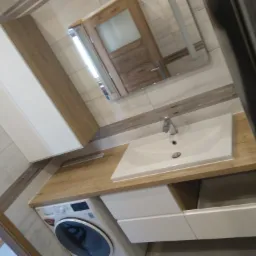 Nowoczesna łazienka z białą umywalką wpuszczoną w drewniany blat, pod którym znajduje się pralka i szafki. Nad umywalką lustro w drewnianej ramie. Ułożenie płytek imituje drewno.