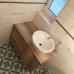 Umywalka nablatowa w okrągłym kształcie z baterią w stylu retro, osadzona na szafce z drewnopodobnym blatem, na tle wzorzystej podłogi i ścian wyłożonych płytkami imitującymi drewno.