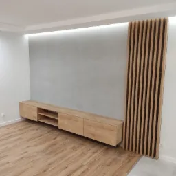 Nowoczesny salon z podwieszaną szafką RTV z jasnego drewna, dekoracyjną ścianą z drewnianych lameli i szarą ścianą z podświetleniem LED, na podłodze panele imitujące drewno.