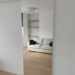 Wnętrze pokoju z przesuwną szafą z lustrzanymi drzwiami, odbijającymi beżową sofę z poduszkami i drewniane półki na białej ścianie.