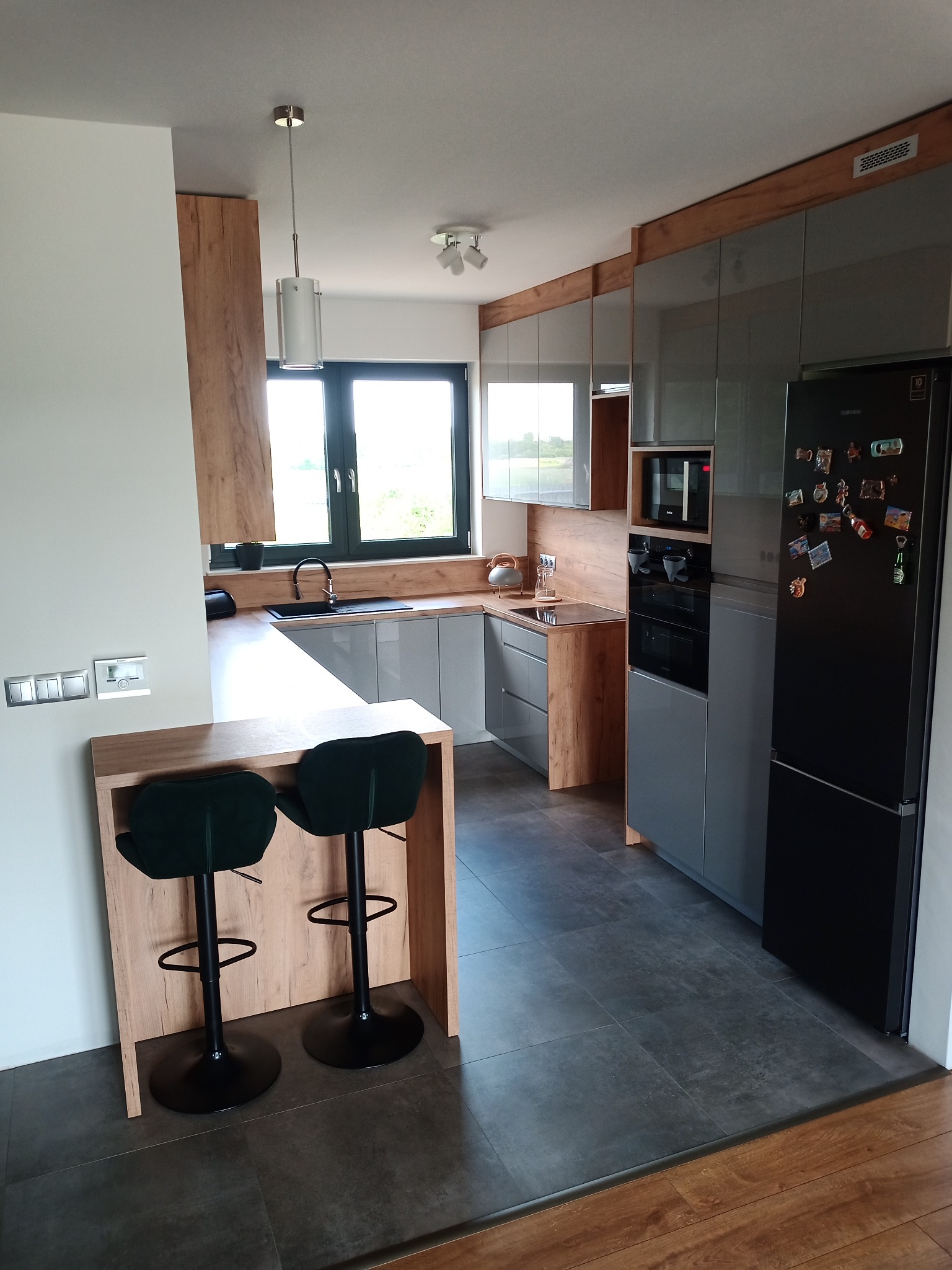 Nowoczesna kuchnia z szarymi szafkami i drewnianymi akcentami, wyspa z dwoma zielonymi hokerami, czarna lodówka z magnesami, okno z widokiem na zieleń.