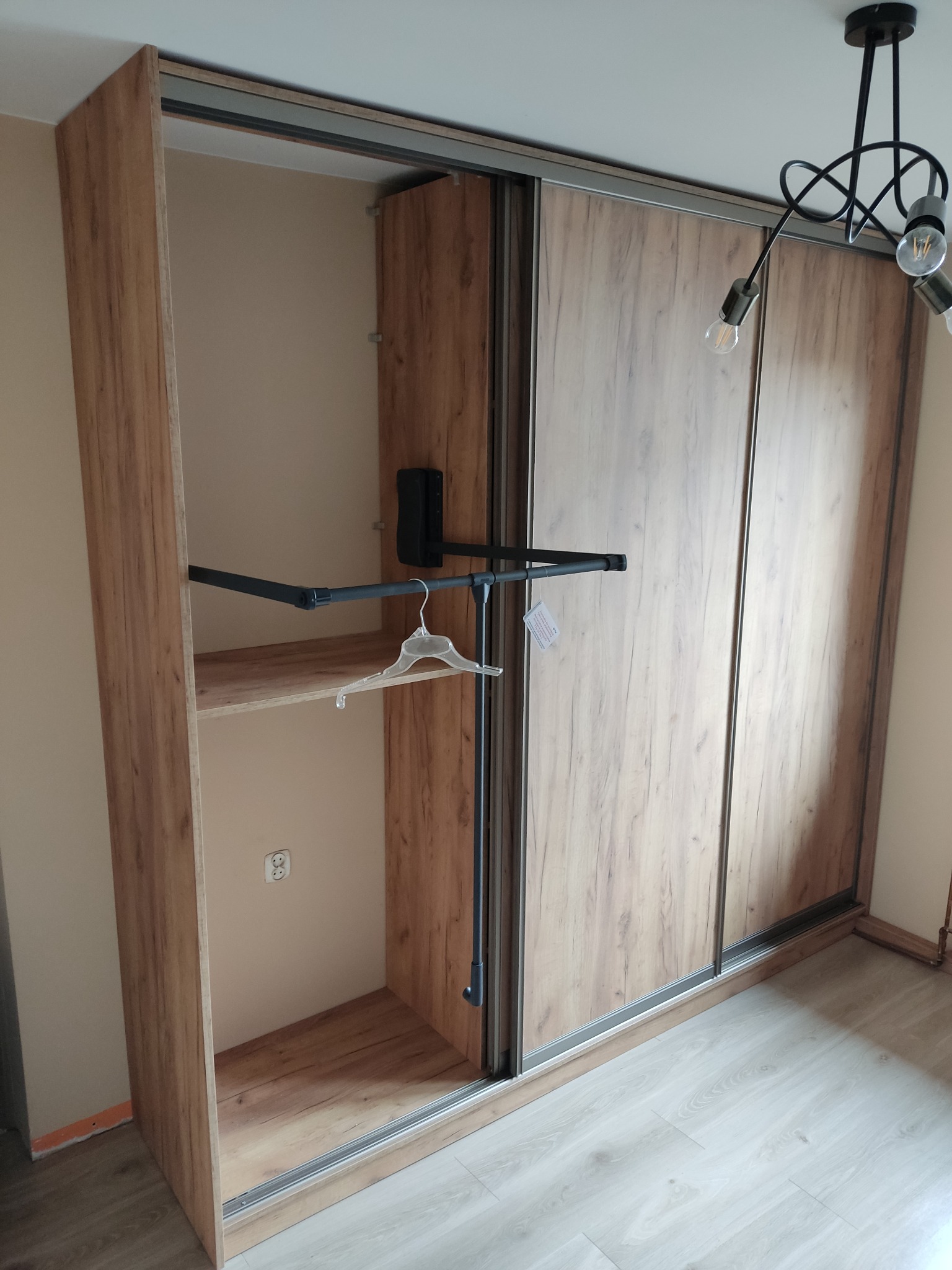 Wnętrze szafy przesuwnej z drewnopodobnymi frontami, widoczne półki, drążek na wieszaki z jednym wieszakiem, oświetlenie sufitowe w tle.