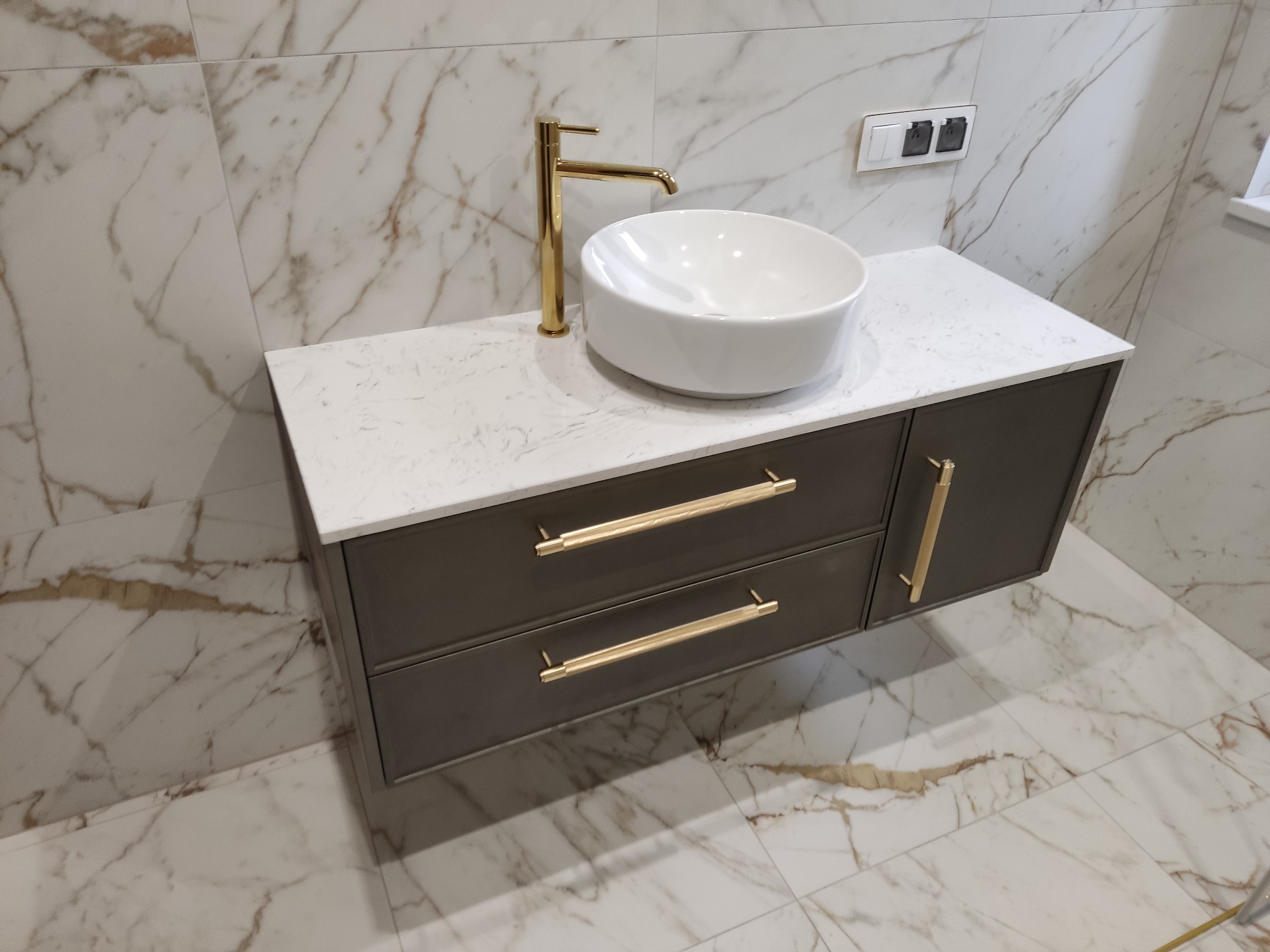 Nowoczesna łazienka z umywalką nablatową, złotą armaturą i szafką z szufladami na tle marmurowych płytek.