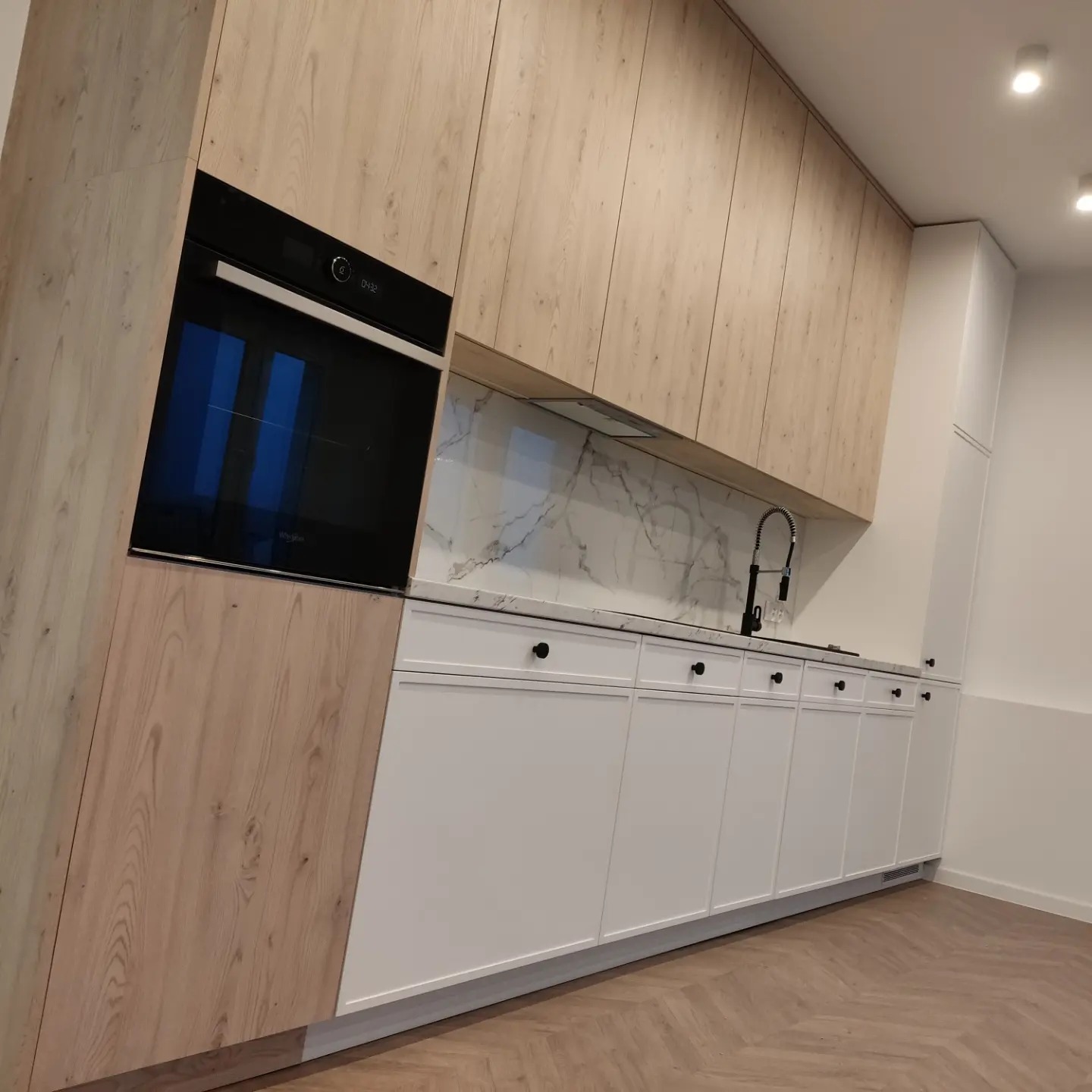 Nowoczesna kuchnia z drewnopodobnymi górnymi szafkami i białymi dolnymi szafkami z czarnymi gałkami, marmurowy backsplash i wbudowany piekarnik.