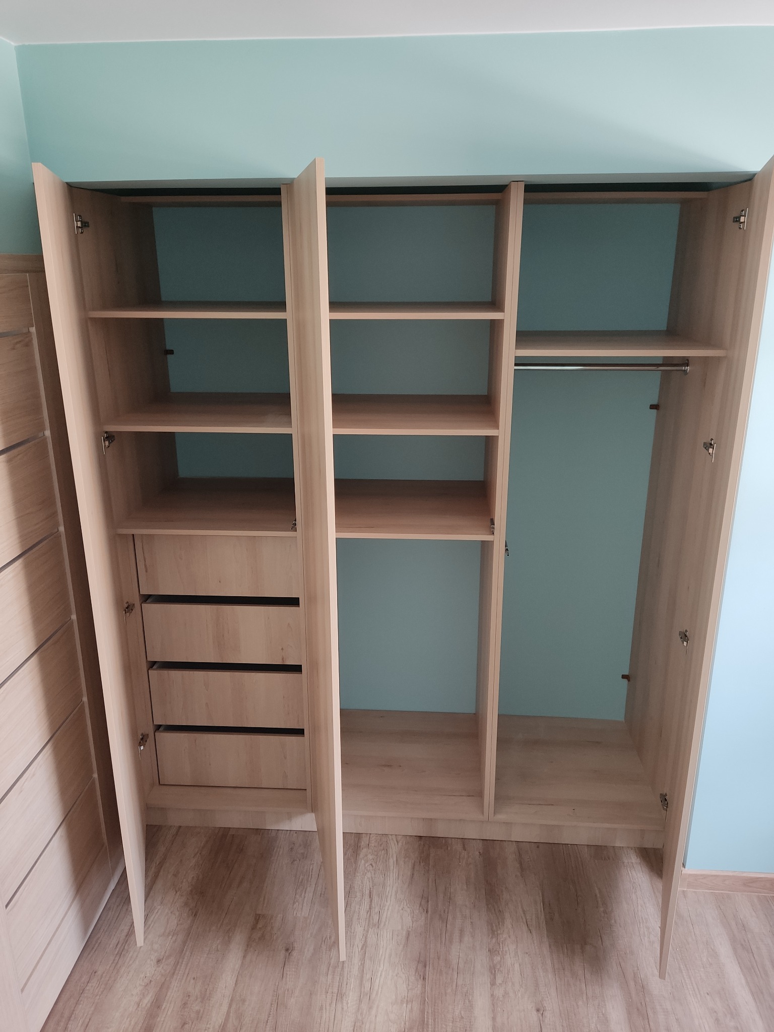 Otwarta, trzydrzwiowa szafa z jasnego drewna z półkami i szufladami, widok na wnętrze szafy na tle błękitnej ściany.