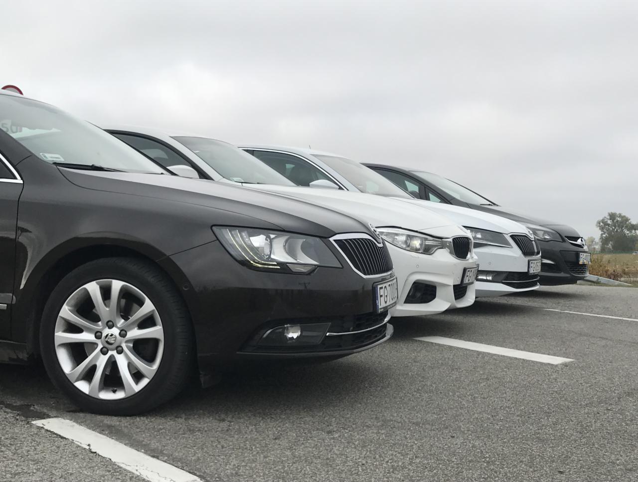 Rząd zaparkowanych samochodów osobowych różnych marek, ustawionych w jednej linii na szarym asfalcie parkingu w pochmurny dzień.