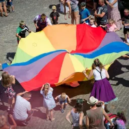 Zabawa z kolorową chustą animacyjną z dziećmi i dorosłymi na brukowanym placu, widok z góry, w tle budynki.