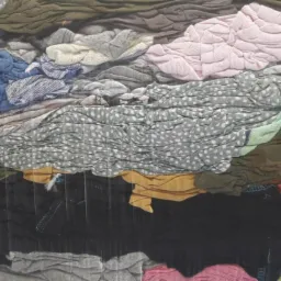 Ściśnięty pakiet różnorodnych tkanin w folii, widoczne wzory w kropki, paski i gładkie faktury w odcieniach szarości, różu, zieleni i pomarańczy.