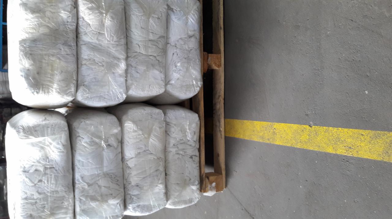 Białe bele tkanin zapakowane w folię na drewnianej palecie, widok z góry, szara betonowa podłoga z żółtą linią w tle.