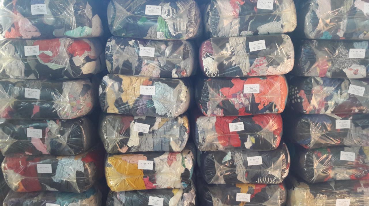 Ułożone w stos paki z odzieżą używaną, zapakowane w przezroczystą folię i opatrzone etykietami 'CZYŚCIWO MIX'.