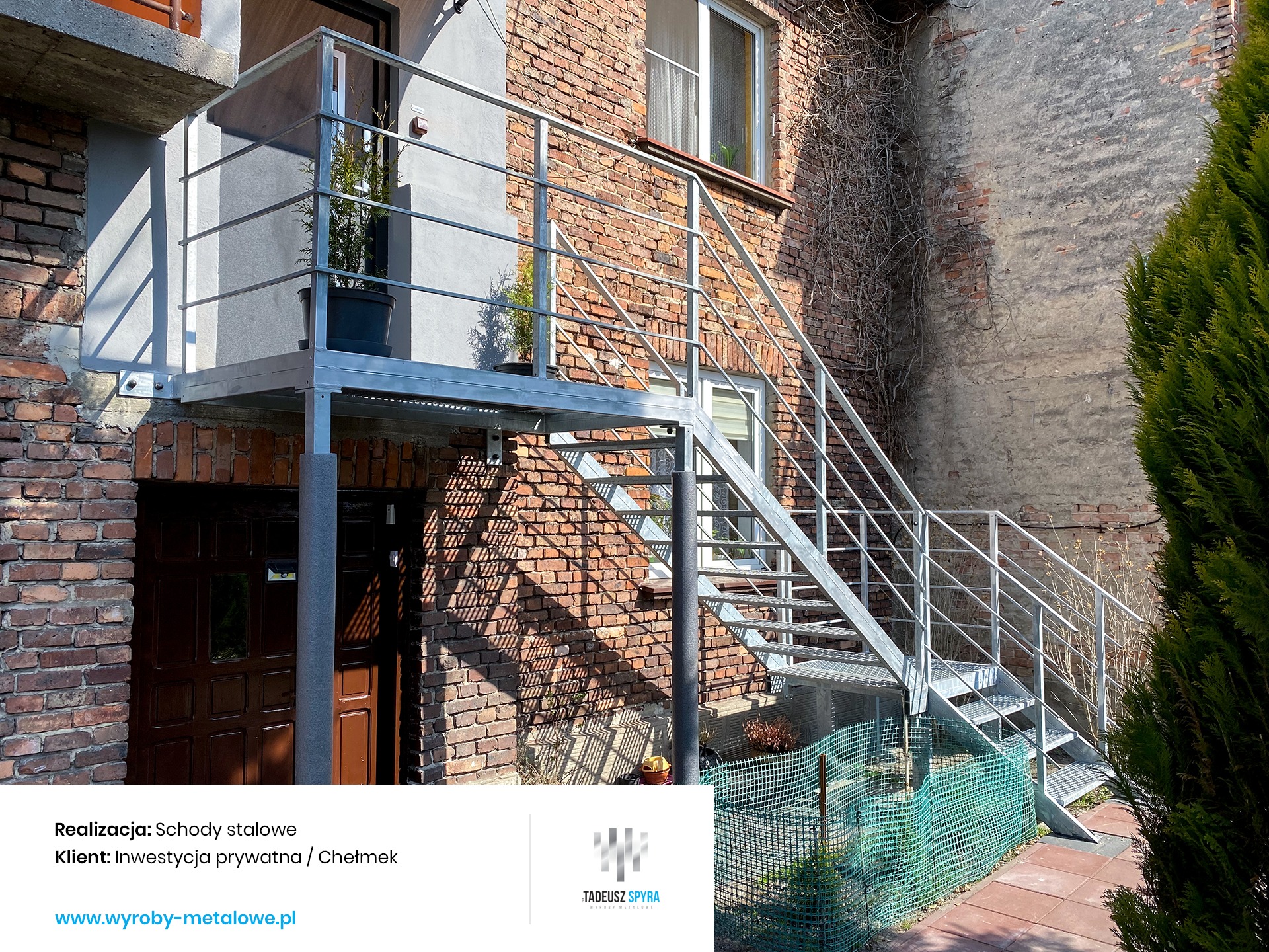 Zewnętrzne schody z metalu prowadzące na balkon, konstrukcja ocynkowana, tło z czerwonej cegły, widoczna zieleń w donicach na balkonie i krzew obok schodów.