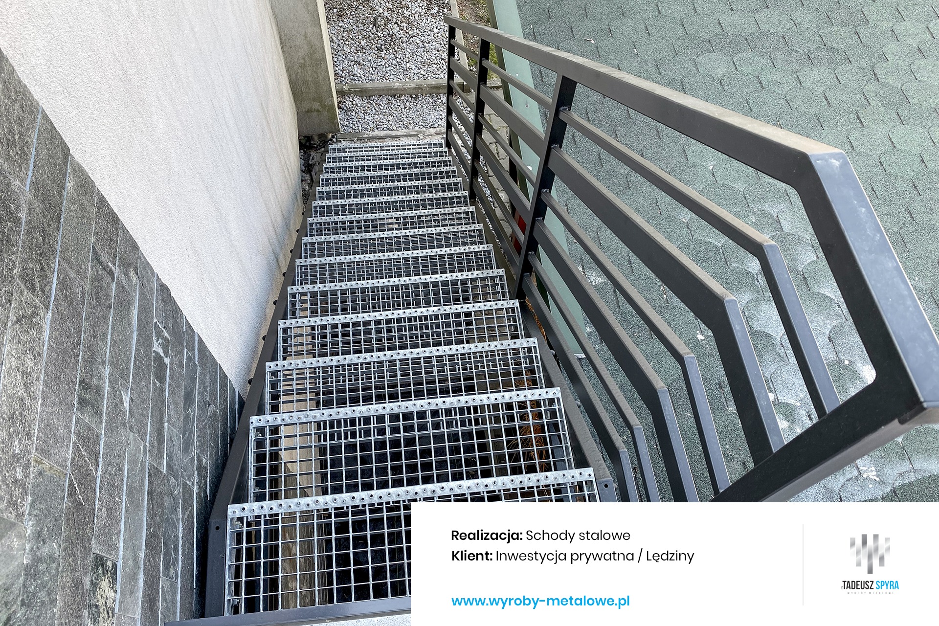 Zewnętrzne schody z metalowej kraty, prowadzące w dół, z szarą poręczą i widocznym fragmentem budynku z kamienną elewacją po lewej stronie.