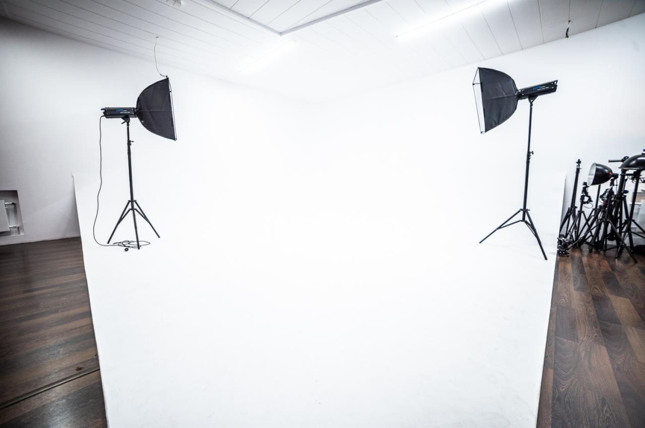 Pusta przestrzeń studyjna z białym tłem, oświetlona przez dwa softboxy na statywach i dodatkowy sprzęt oświetleniowy ustawiony z boku na drewnianej podłodze.