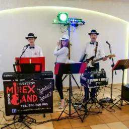 Trzyosobowy zespół w jednakowych kapeluszach i koszulach z muszkami, grający muzykę na weselu lub innej uroczystości, z widocznym banerem reklamowym zespołu na statywie.