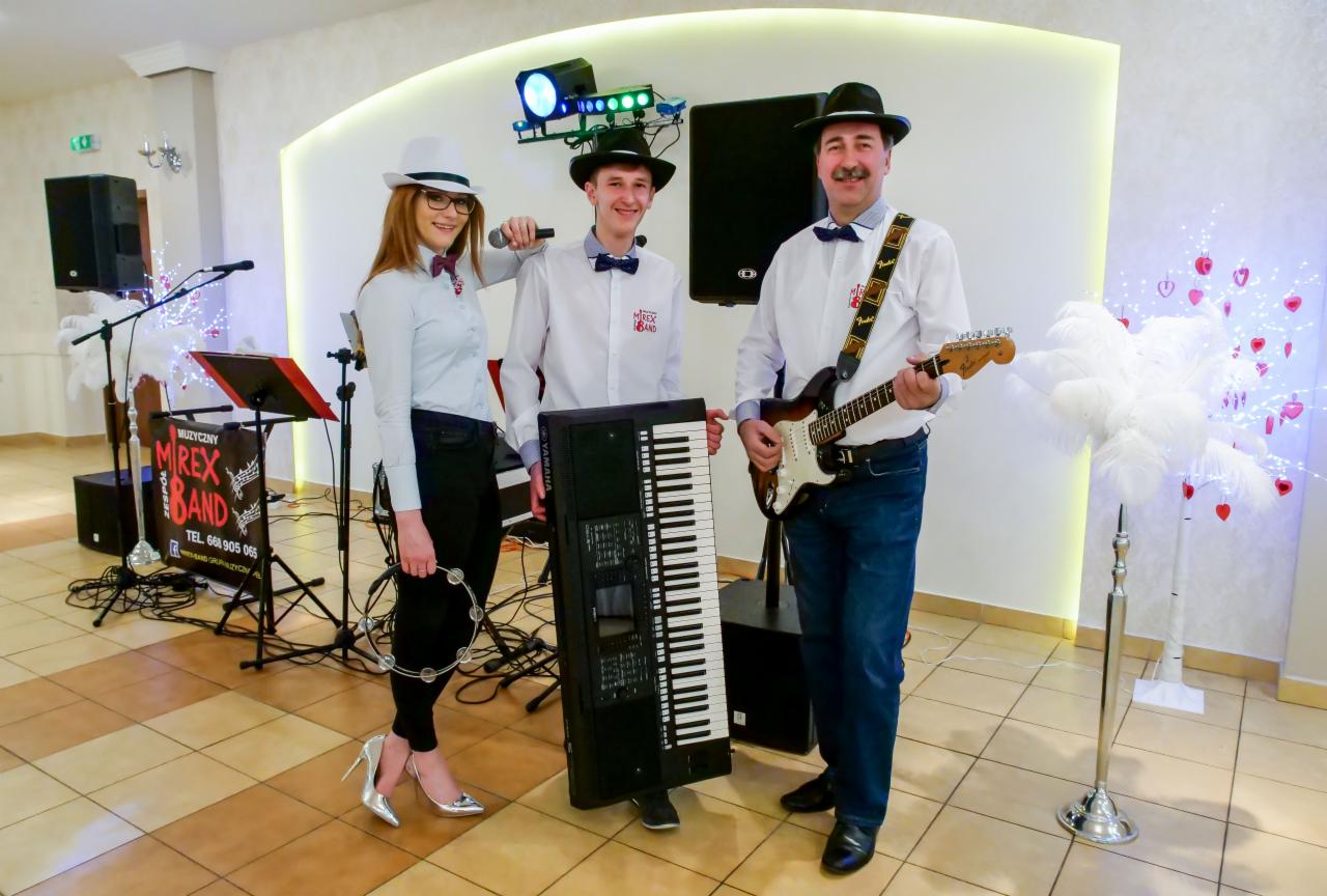 Trzyosobowy zespół w kapeluszach prezentuje się z gitarą, keyboardem i tamburynem na tle sali z dekoracjami.
