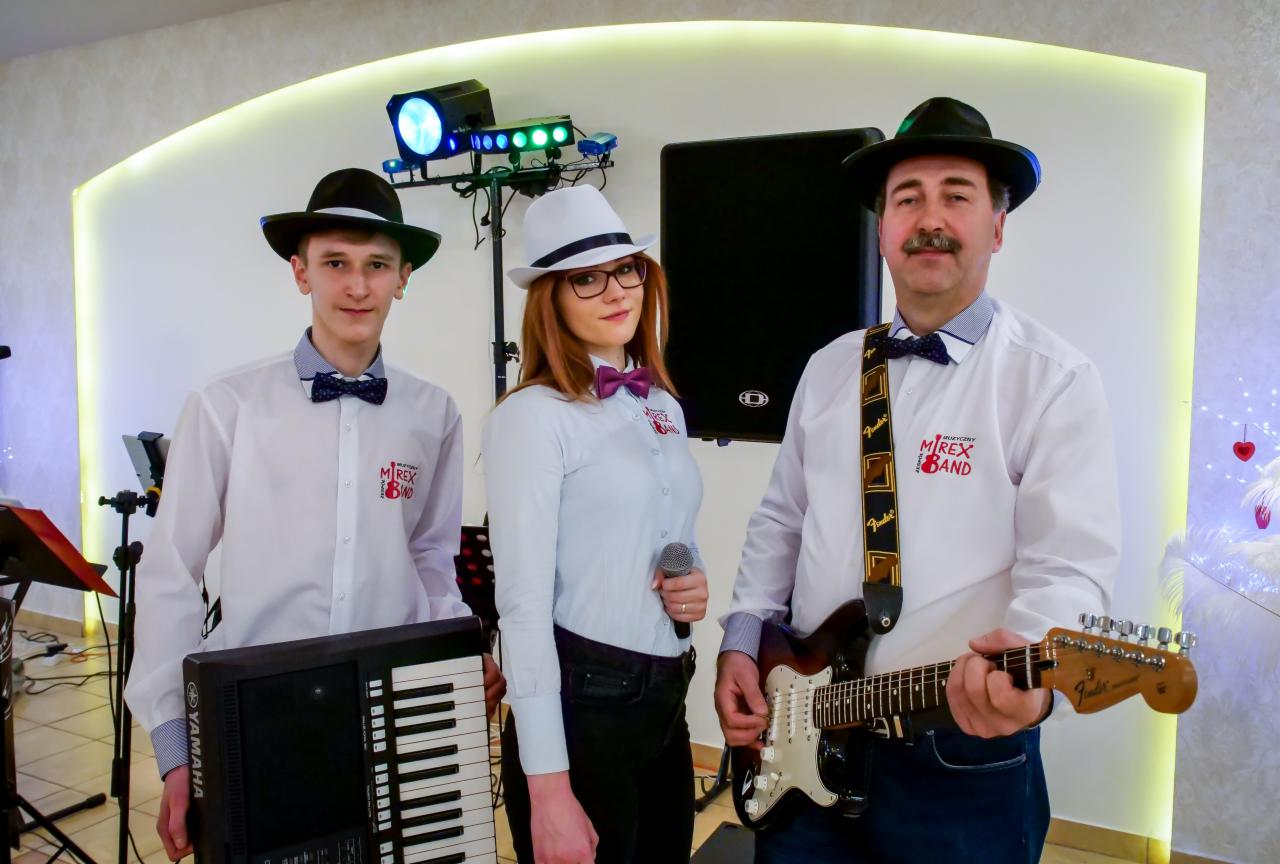 Trzyosobowy zespół w białych koszulach z muszkami i kapeluszach, mężczyzna z gitarą Fender, kobieta z mikrofonem, młody mężczyzna z keyboardem Yamaha, logo zespołu na koszulach, oświetlenie...