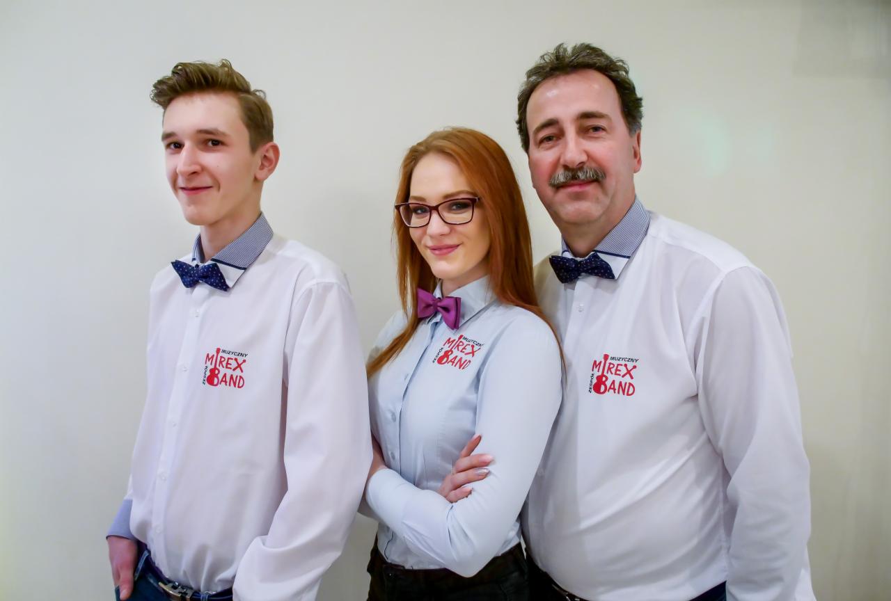 Trzyosobowy skład w koszulach z logo zespołu i muchami, pozujący na tle jasnej ściany.