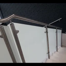 Nowoczesna balustrada schodowa ze stali nierdzewnej i białego szkła, widok z góry na schody z czarną ścianą z cegły w tle.