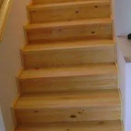 Proste, drewniane schody z jasnego drewna, widok z dołu, z widoczną poręczą po lewej stronie i fragmentem ściany w tle.