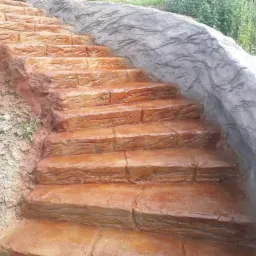 Betonowe schody zewnętrzne imitujące kamień, z nier&oacute;wną fakturą i brązowo-pomarańczowym wybarwieniem, prowadzące w g&oacute;rę zbocza porośniętego trawą, z szarą ścianą oporową po prawej stronie.