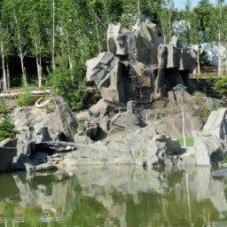 Sztuczny wodospad z szarego kamienia kaskadowo opadający do zielonkawego stawu w ogrodzie w Bydgoszczy, otoczony drzewami i krzewami.