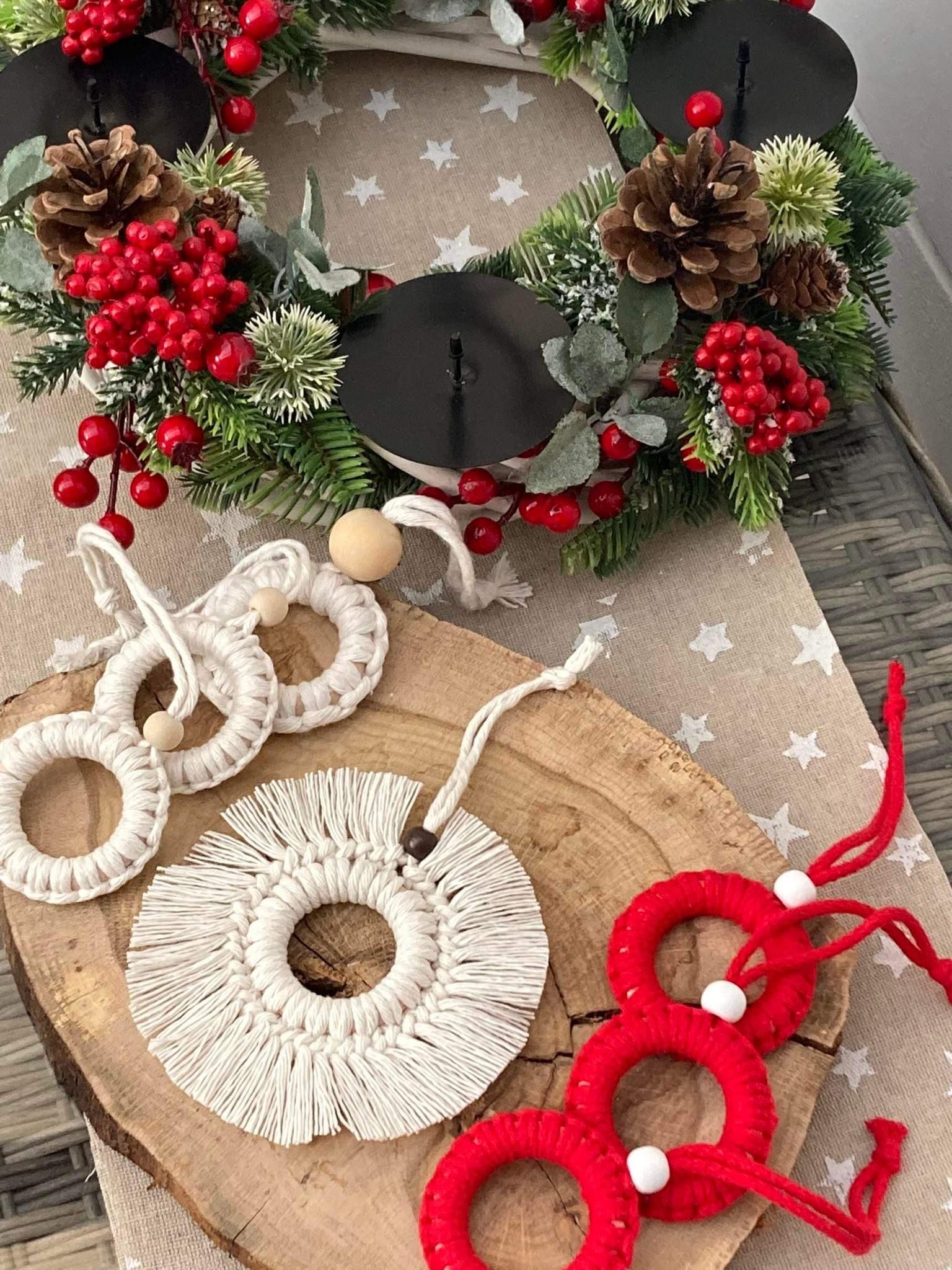 Dekoracyjny wieniec adwentowy z czarnymi świecznikami, szyszkami i czerwonymi jagodami, obok ręcznie robione, białe i czerwone ozdoby choinkowe z makramy i włóczki, ułożone na drewnianym plastrze.