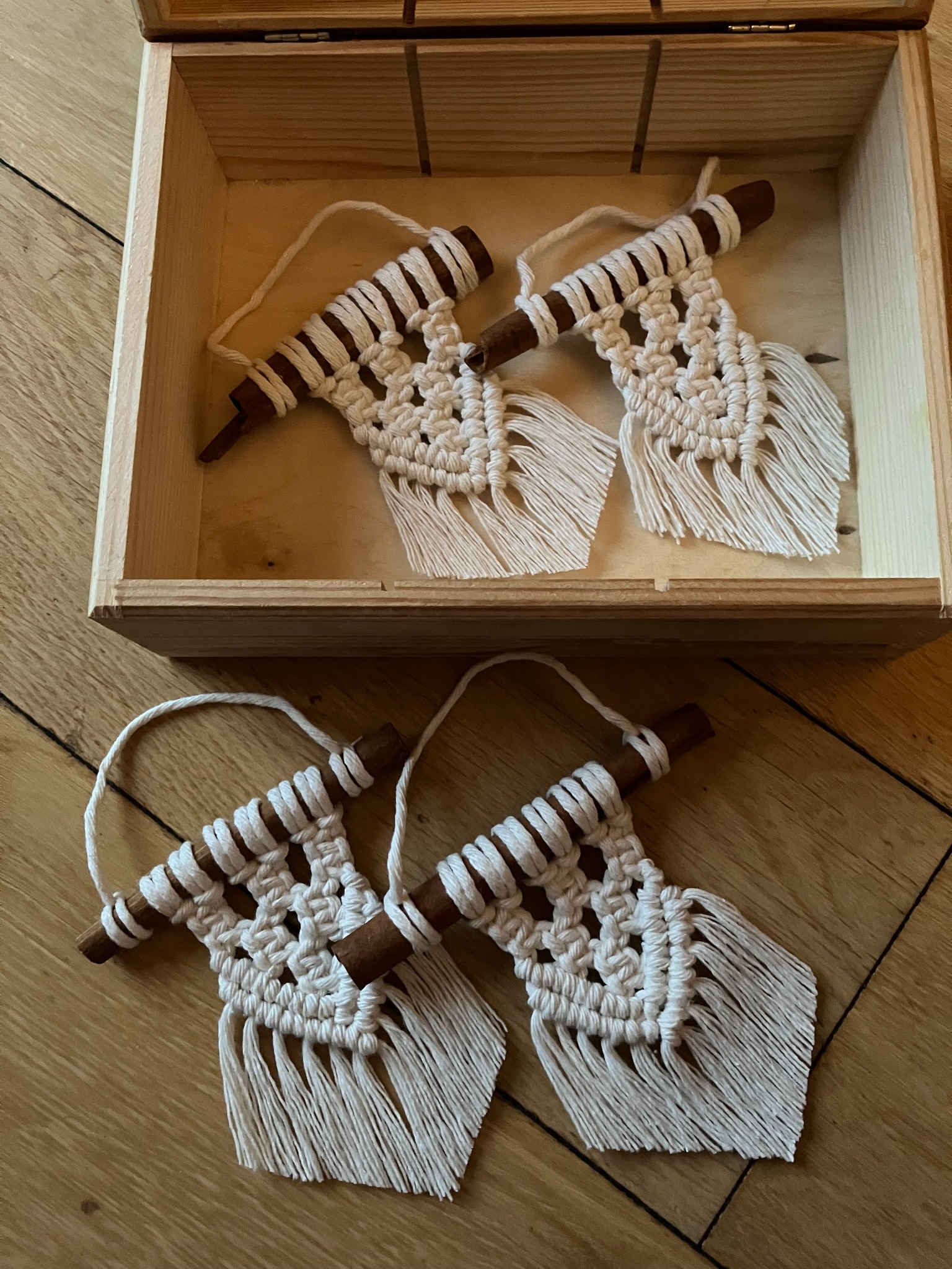 Ręcznie robione, białe makramy z drewnianymi poprzeczkami, część w drewnianej skrzynce, na drewnianej podłodze, widok z góry.