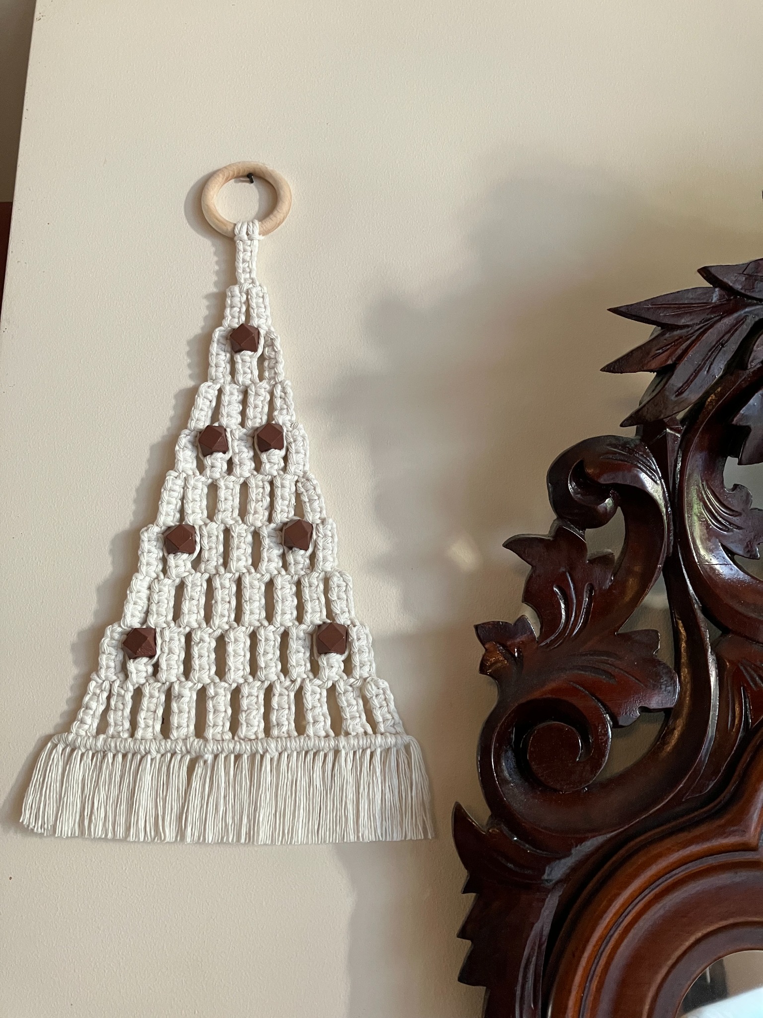 Ręcznie robiona makrama w kształcie choinki, zdobiona brązowymi koralikami, zawieszona na drewnianym kółku, obok fragmentu ciemnego, rzeźbionego lustra.