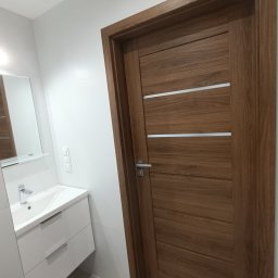 Usługi Wykończeniowo-Remontowe Marcin Świtalski - Nowoczesna łazienka z drewnianymi drzwiami z trzema srebrnymi intarsjami, białą umywalką z szafką i lustrem na białej ścianie wyłożonej płytkami.