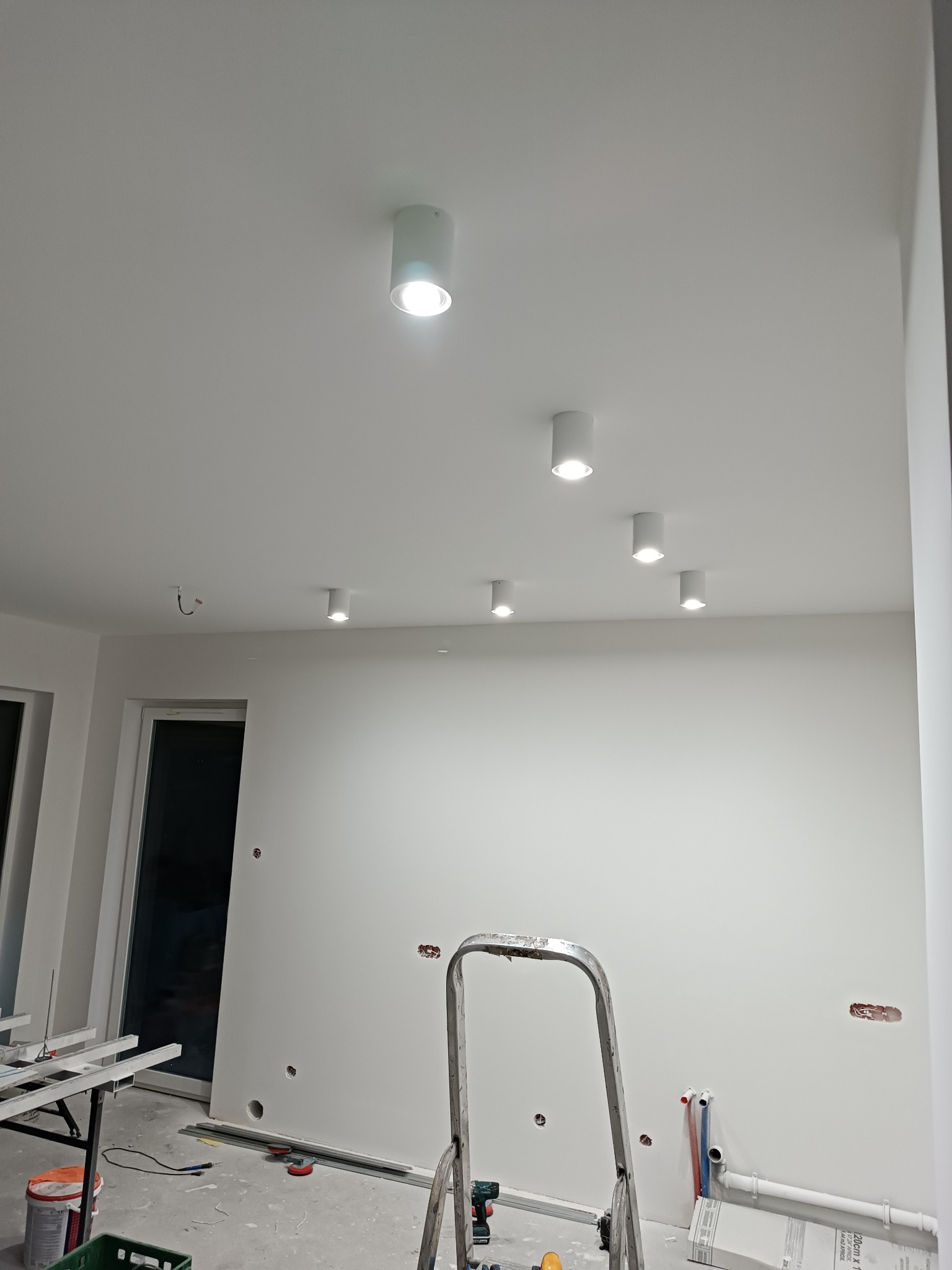 Białe wnętrze w trakcie remontu: zamontowane reflektory sufitowe, ślady po gładzi, drabina, profile i narzędzia na podłodze.