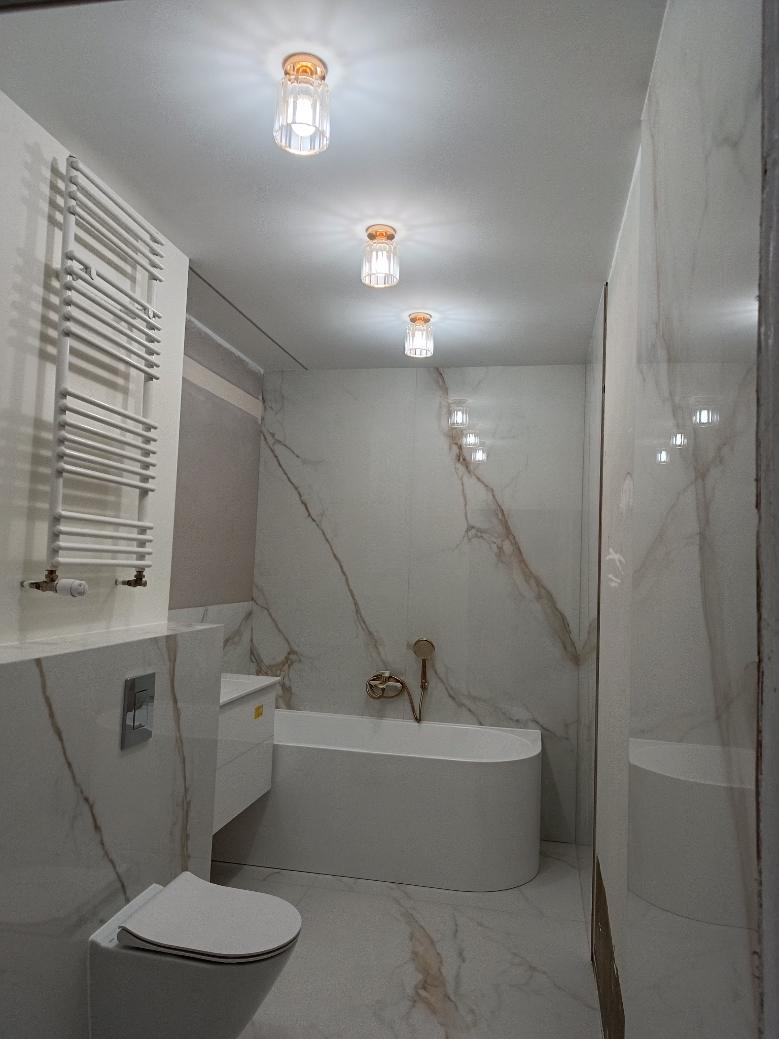 Nowoczesna łazienka z marmurowymi płytkami, wiszącą toaletą, grzejnikiem drabinkowym i podłużną wanną z złotą armaturą, oświetlona trzema kryształowymi lampami sufitowymi.