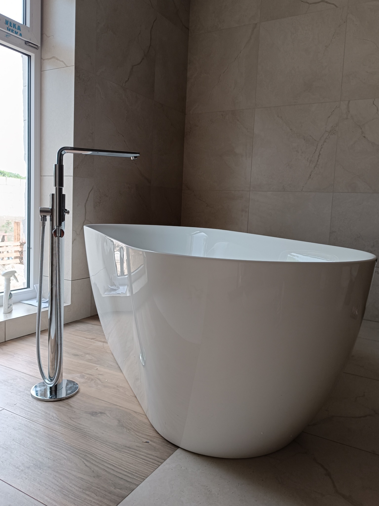 Wolnostojąca biała wanna z chromowaną baterią podłogową w nowoczesnej łazience wykończonej płytkami w odcieniach beżu i szarości, widok na okno z zewnątrz.