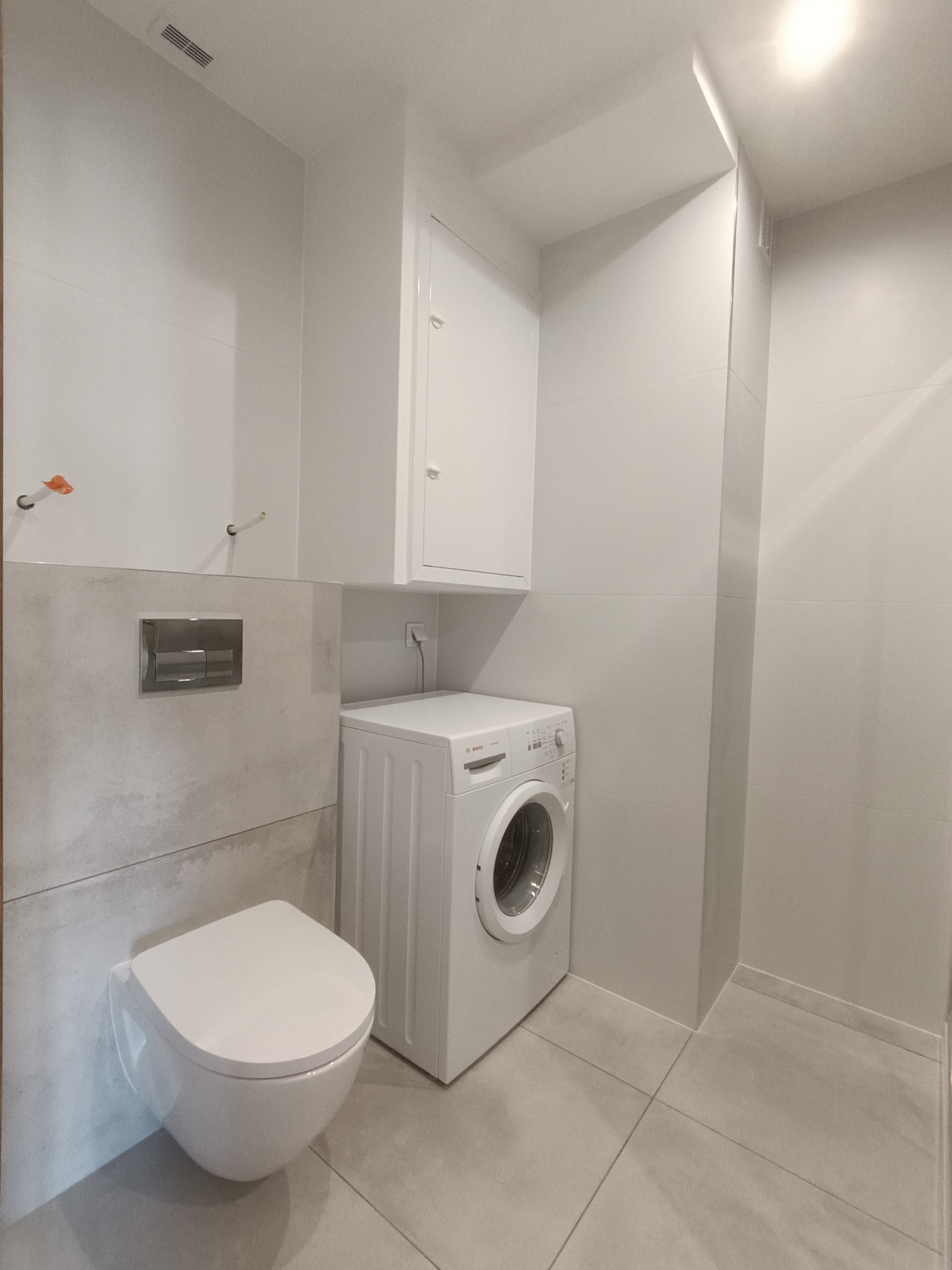 Nowoczesna łazienka w Lubaszu po remoncie, z podwieszaną toaletą, pralką i szarą glazurą, widoczne niedokończone podłączenia.