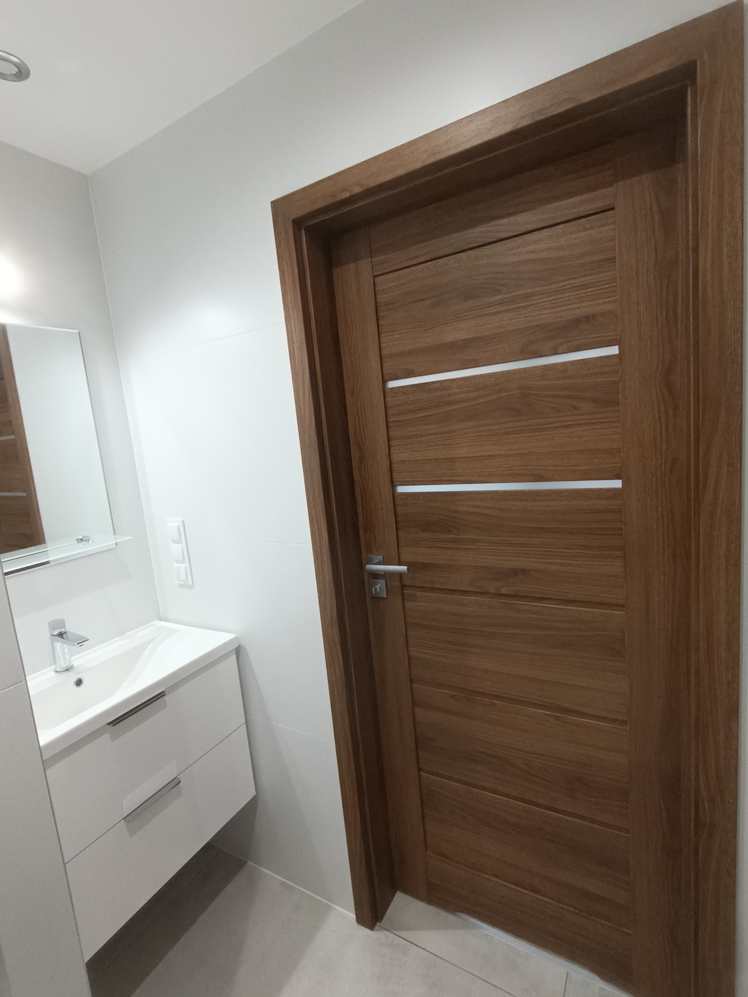 Nowoczesna łazienka z drewnianymi drzwiami z trzema srebrnymi intarsjami, białą umywalką z szafką i lustrem na białej ścianie wyłożonej płytkami.