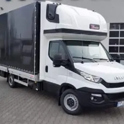 Biały samochód dostawczy Iveco z czarną plandeką na tle szarego budynku z numerem 11 nad bramą.