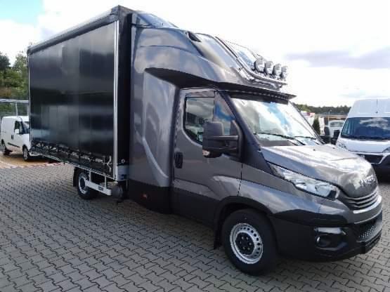 Szary samochód dostawczy z plandeką, dodatkowym oświetleniem na dachu i małym białym busem w tle, stojący na kostce brukowej.