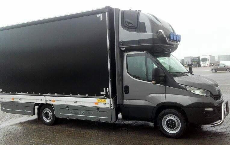 Szary samochód dostawczy z czarną plandeką i dodatkowym modułem nad kabiną kierowcy, widoczne zabudowania przemysłowe w tle.