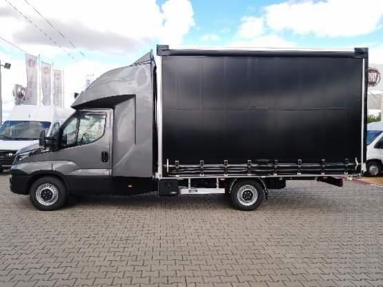 Szary samochód dostawczy z czarną plandeką na parkingu z kostki brukowej, widok z boku.