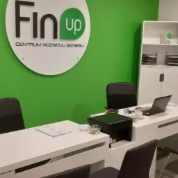 Nowoczesne biuro firmy finansowej z logo FinUp na zielonej ścianie, białe biurka z laptopami i krzesłami dla klientów, schludne i profesjonalne wnętrze.