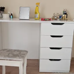 Biała toaletka z czterema szufladami, lustrem z podświetleniem LED, pędzle do makijażu w złotym pojemniku, flakoniki perfum na złotej tacy i stołek z kwiatowym wzorem.