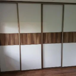 Czterodrzwiowa szafa wnękowa z przesuwnymi drzwiami, białe panele z poziomym pasem dekoracyjnym w kolorze drewna, widoczna podłoga z paneli imitujących drewno.