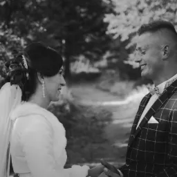 Czarno-białe zdjęcie uśmiechniętej pary młodej trzymającej się za ręce na tle leśnej ścieżki, panna młoda w welonie i futerku, pan młody w kraciastej marynarce i muszce.